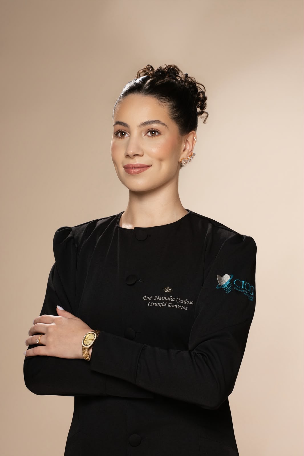 Dra. Nathália Cardoso de jaleco preto, braços cruzados, sorrindo levemente em fundo bege